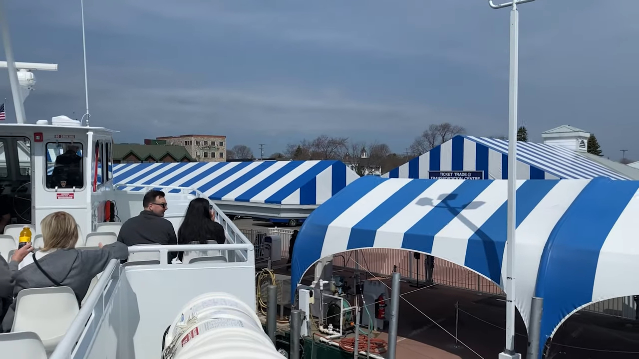 Passengers on a ferry deck near docks with blue-and-white striped awnings, preparing to depart for Mackinac Island