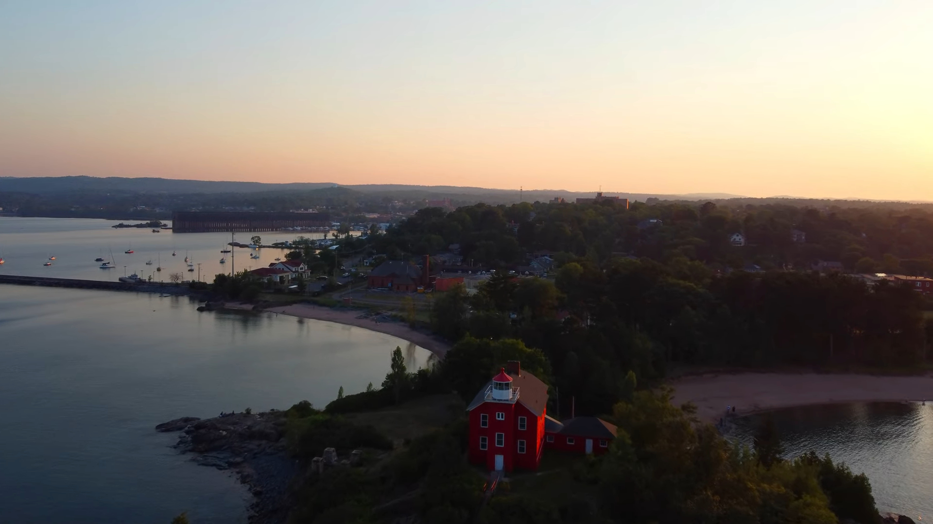 Aerial view of Marquette Harbor Lighthouse, a bright red building near the shoreline, with a calm bay and hills in the background at sunset