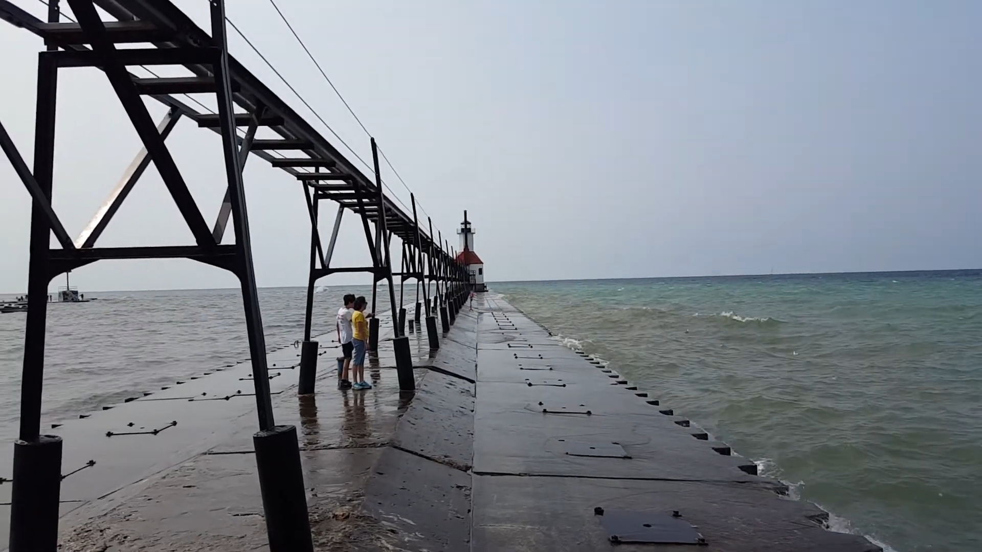 People walking along the pier towards St. Joseph North Pier Lighthouse, with metal catwalks above and waves crashing on the sides