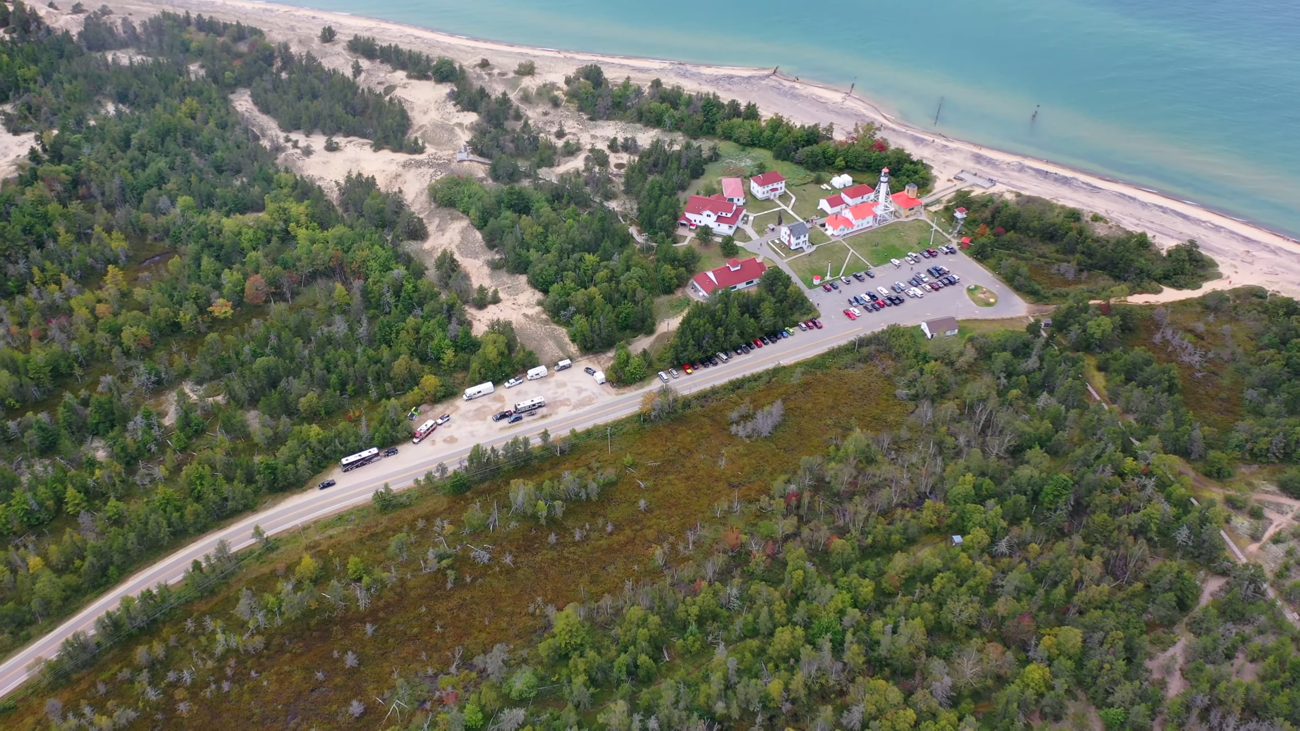 Aerial view of Whitefish Point Light Station, featuring red-roofed buildings surrounded by dense greenery, sandy dunes, and the shoreline of Lake Superior