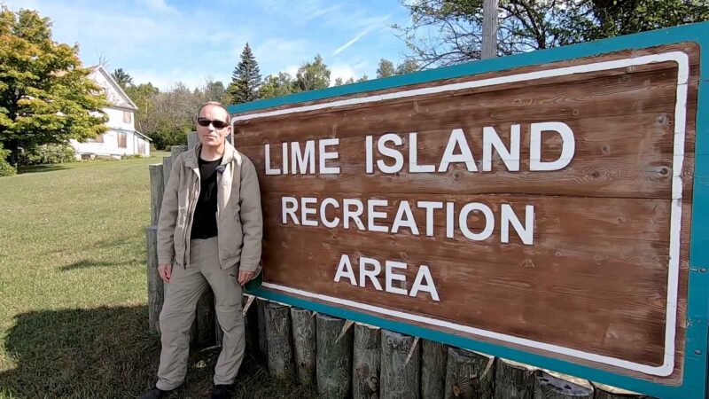 Person standing beside a wooden sign reading “Lime Island Recreation Area” on a grassy area with trees and an old white building in the background, Upper Michigan